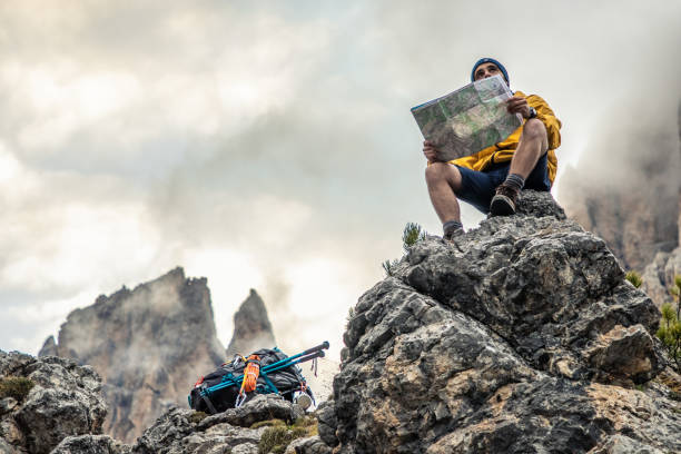 joven excursionista sentado en el mapa de lectura de montaña de piedra, con cielo nublado y niebla. chaqueta amarilla, mochila, barba negra y gorro. dolomitas itinerantes, italia. - montañas dolomita fotografías e imágenes de stock