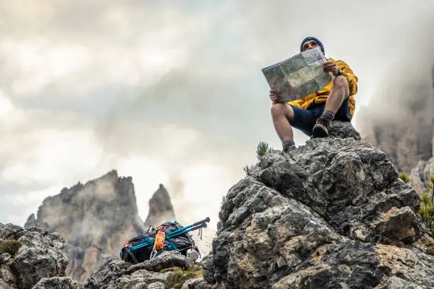 Photo of Young man hiker sitting on stone mountain reading map, with cloudy sky and fog. Yellow jacket, backpack, black beard and beanie. Traveling dolomites, Italy.