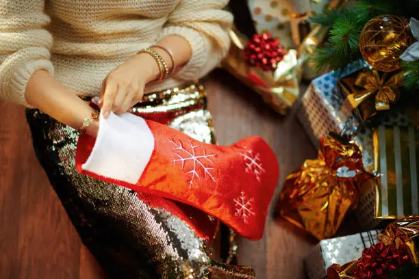 Closeup on young woman in gold sequin skirt and white sweater under decorated Christmas tree near present boxes takes something out of red Christmas stocking.