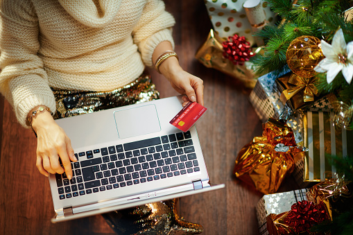 trendy housewife in gold sequin skirt and white sweater with credit card making online shopping on e-commerce site and under decorated Christmas tree near present boxes.