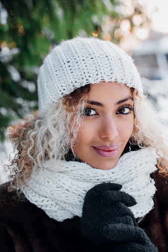 Portrait of beautiful young black woman wearing in fur coat and knitted hat walking in winter park