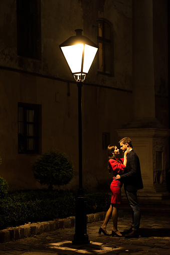 young, elegant couple hugging while standing under street lamp at night