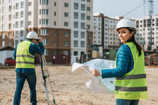 istock Selective focus of surveyor with blueprint smiling at camera and colleague using digital level on construction site 1193073998