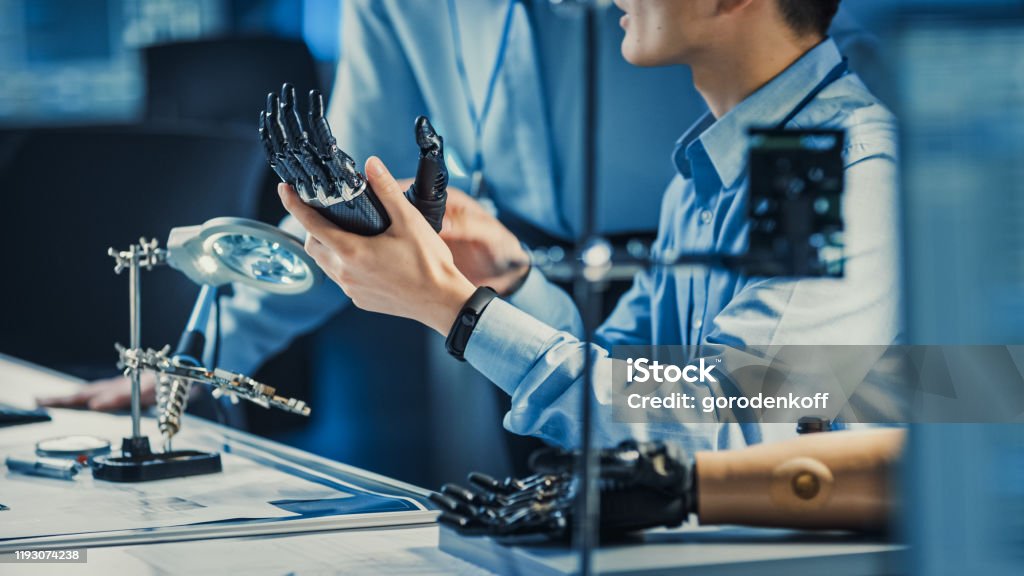 Technological Prosthetic Robot Arm is Tested by Two Professional Development Engineers in a High Tech Research Laboratory with Modern Futuristic Equipment. Compare Data on a Personal Computer. Healthcare And Medicine Stock Photo