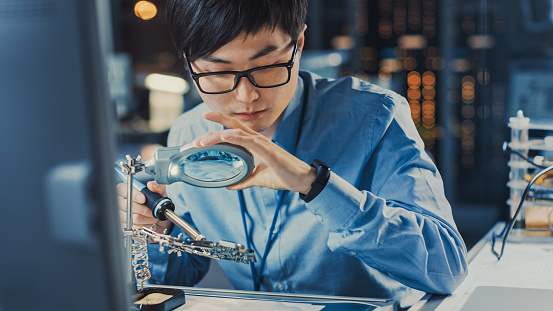 Close Up of a Professional Japanese Electronics Development Engineer in Blue Shirt Soldering a Circuit Board in a High Tech Research Laboratory with Modern Computer Equipment.