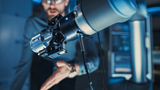 Close Up of a Futuristic Robotic Arm Moving a Metal Detail. Team of Engineers Discuss This Technologically Advanced Process. They are in a High Tech Research Laboratory with Modern Equipment.