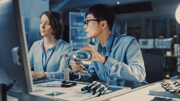 青いシャツを着た日本の開発エンジニアは、現代機器を備えたハイテク研究所の回路基板をはんだ付けしています。彼の同僚は彼にコンプター画面上のクエストオとポイントを尋ねます。 - reference point ストックフォトと画像