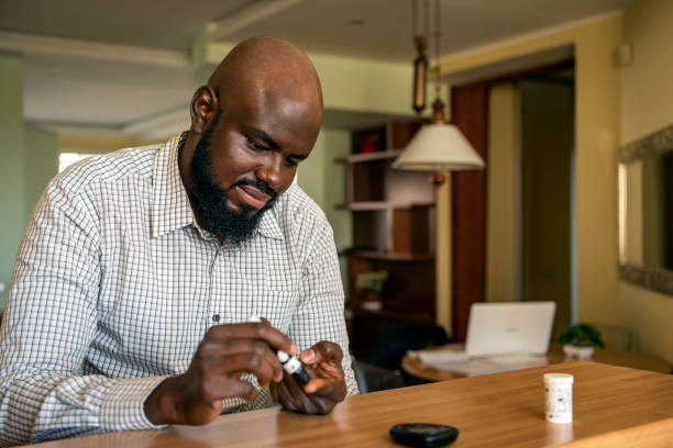 African Businessman Doing Blood Sugar Test at Home A modern African businessman is sitting at the desk at the home office and taking blood from his finger due to diabetes. The daily life of a man of African-American ethnicity person with a chronic illness who is using glucose tester. blood sugar test stock pictures, royalty-free photos & images