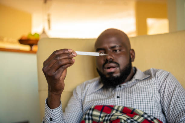 A mature African-American is sitting on the sofa at home in a bad health condition and looking at the thermometer and checking the temperature of his body. An Ill African Man with seasonal infections, flu, allergy lying down in bed, covered with a blanket with high fever and flu.