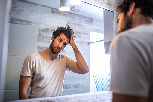 Un joven guapo tocándose el pelo con la mano y acicalándose en el baño en casa. Hombre metrosexual blanco preocupado por la pérdida de cabello y mirando espejo su línea de pelo en retroceso. photo
