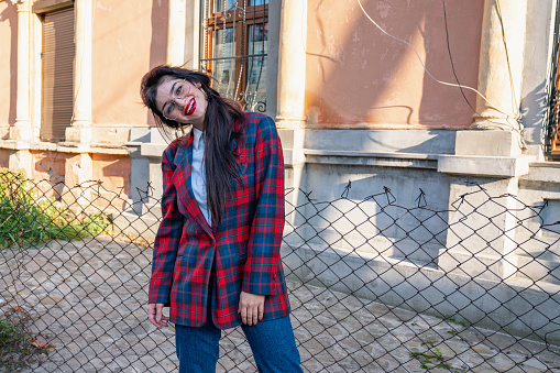 Attaractive brunette girl in glasses dressed in plaid jacket and white shirt bowed her head on left shoulder and looking at the camera stands near the old house, autumn sunny day