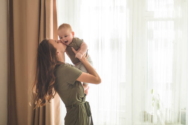 Home portrait of a baby boy with mother near the window. Mom holding and kissing her child. Home portrait of a baby boy with mother near the window. Mom holding and kissing her child. Mother day concept cute boy show love symbol hand stock pictures, royalty-free photos & images