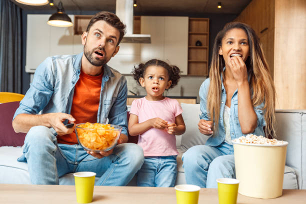 Family watching tv, eating chips and spending day together at home Shocked young adult father and mother sitting on comfort couch with cute african child girl, watching tv, making surprised faces. Family eating chips and spending day together at home family eating potato chips stock pictures, royalty-free photos & images