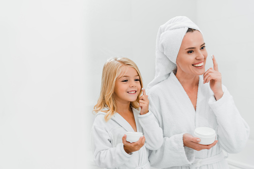 cheerful mother and daughter applying cosmetic cream in bathroom