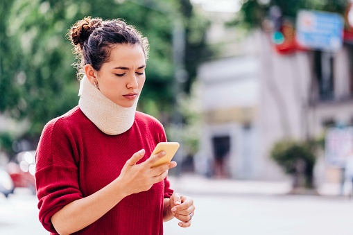 Serious woman text messaging through smart phone. Young female with physical injury is wearing neck brace. She is in red sweater.