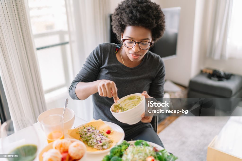Young woman eating vegan food at home A young woman is eating vegan food at home  with ingredients of tomato, avocado paste food, onion, lettuce, apples, broccolis and also a cup of orange juice. Eating Stock Photo