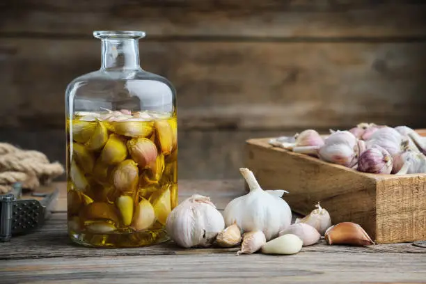 Photo of Garlic aromatic flavored oil or infusion bottle, wooden crate of garlic cloves and  garlic press on wooden kitchen table.