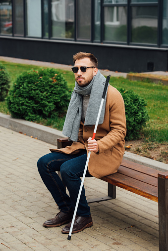 Blind man in coat holding walking stick while sitting on bench