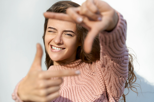 Girl showing frame sign with their hands. Girls Hands taking focus frame shooting like selfie on mobile camera. Portrait of young woman with brown hair, green eyes wearing pink sweater in Park