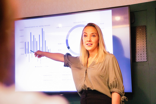 Young blond female manager explaining quarterly results on large led screen with a nice energy. Over the shoulder view suggesting she is addressing someone in particular.