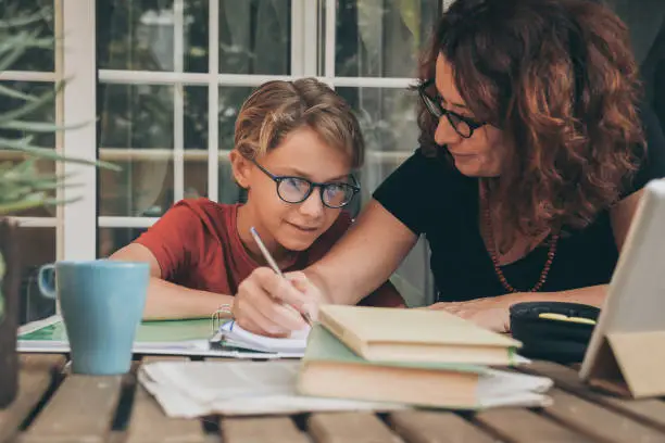 Young student doing homework at home with school books, newspaper and digital pad helped by his mother. Mum writing on the copybook teaching his son. Education, family lifestyle, homeschooling concept