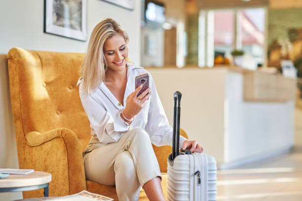 businesswoman with luggage in modern hotel lobby using smartphone - guest imagens e fotografias de stock