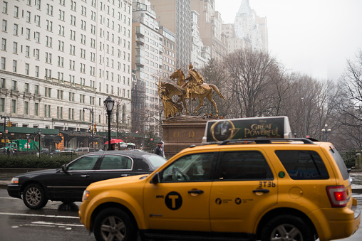 New York City, USA - February 27, 2013: It is raining in New York City. Most people on Fifth Avenue (and Central Park South) have an umbrella to keep them dry. Some cars, including yellow taxi cabs, are driving southbound. In the background is the Grand Army Plaza with the William Tecumseh Sherman monument (or Sherman Memorial or Sherman Monument).
