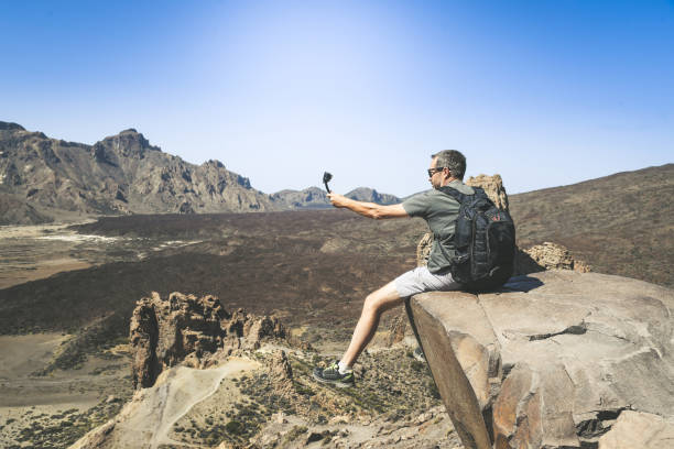 istock Sporty man with backpack making video with action cam to sharing online. Caucasian boy taking picture sitting on a mountain cliff overlooking the panorama. Holiday trip travel, new trend tech concept. 1193142825