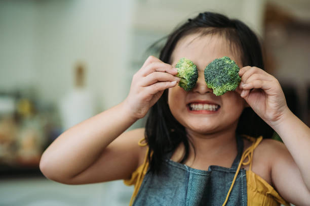 la bambina asiatica cinese si comporta carina con la mano che tiene i broccoli che le mettono davanti agli occhi con il viso sorridente in cucina - childrens food foto e immagini stock