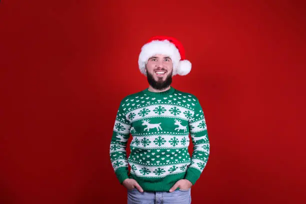 Studio portrait of handsome bearded man wearing christmas sweater with snowflake ornament, posing over the red wall, copy space for text. Festive background. Male with facial hair smiling.