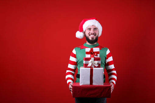 Studio portrait of handsome bearded man wearing traditional elf costume, green vest & striped sleeve, posing over the red wall, copy space for text. Festive background. Male with facial hair smiling.