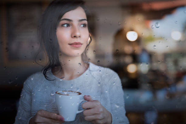 jeune femme restant devant la fenêtre et appréciant une tasse de café à la maison un jour pluvieux - women thinking contemplation sideways glance photos et images de collection