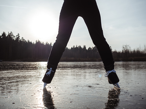 Legs of a girl in ice skates spread shoulder-width apart on the ice of a lake against a forest.