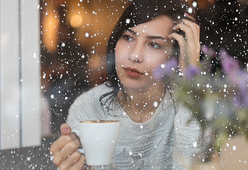Beautiful Young Woman drinking coffee and watching snow through the window