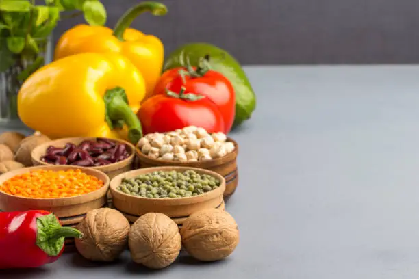 Photo of A set of products for diet food. On a gray background, walnuts, chickpeas, beans, lentils, yellow and red peppers, tomatoes, avocado. There is a place for text