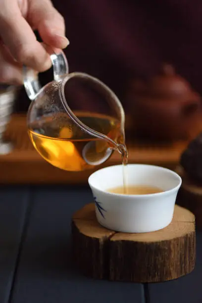 Healthy beverage concept. Pouring freshly brewed tea from glass pitcher into porcelain cup on wood slab on dark background copy space