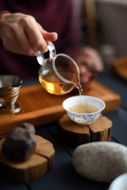 Tea ceremony. Slender woman hand pouring freshly brewed green tea from glass pitcher into Chinese cup and decorative stones on wood slabs on dark background closeup