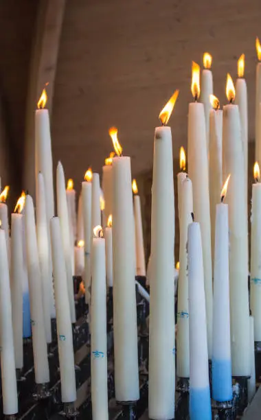 Photo of Big burning candles from prayers for hope. Saint Bernadette grotto with many white candles with flame. Sanctuary in Lourdes. Melting wax and candlelight.