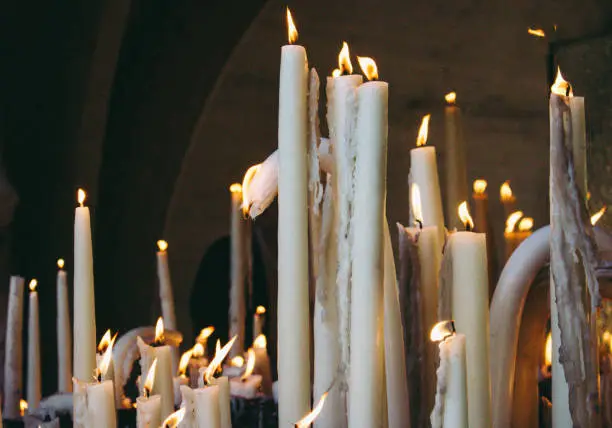 Photo of Big burning candles from prayers for hope. Saint Bernadette grotto with many white candles with flame. Sanctuary in Lourdes. Melting wax and candlelight.
