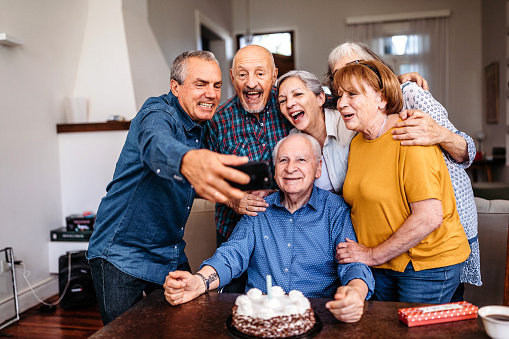 Cheerful friends taking selfie using mobile phone camera while celebrating birthday of senior man at nursing home