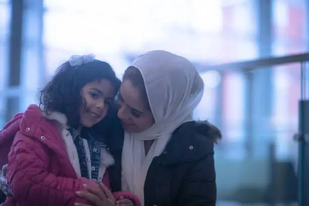 A Muslim mother sits with her daughter in the airport waiting. They are both wearing coats and dressed casually and the mother has Hijab on.  The little girl is sitting on her mothers lap and they have their heads nestled in close to one another.  The mother is looking down at her daughter and smiling.