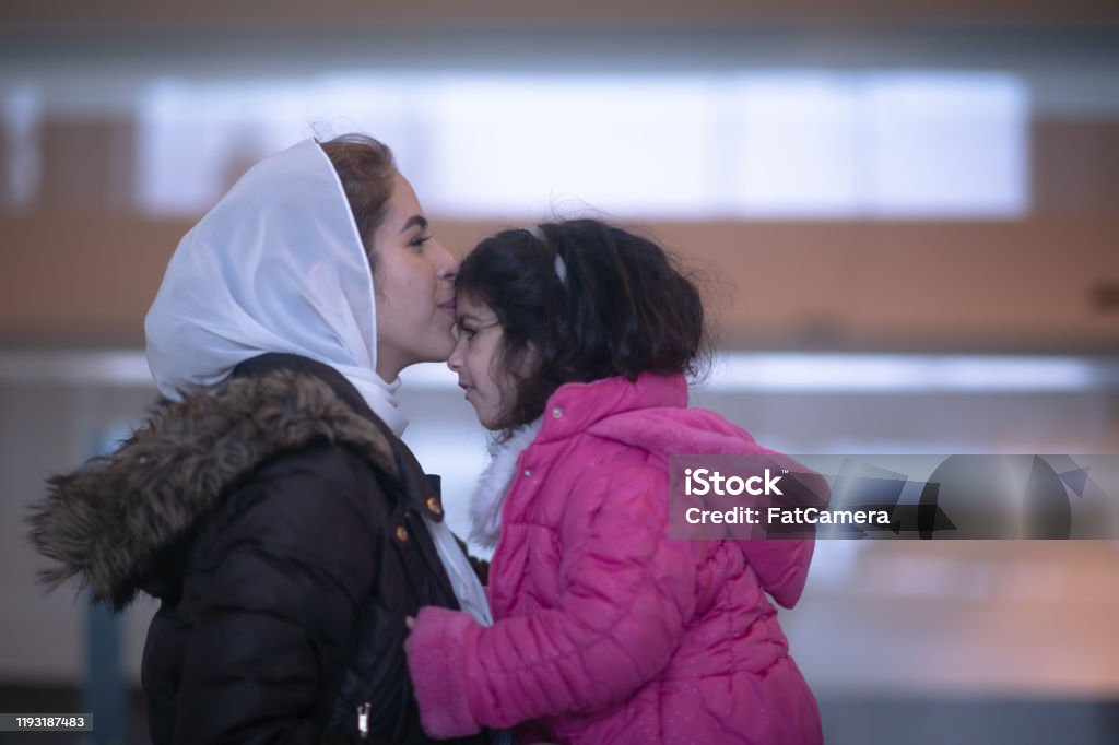 Muslim Mother and Daughter in the Airport stock photo A Muslim mother sits with her daughter in the airport waiting. They are both wearing coats and dressed casually and the mother has Hijab on.  The little girl is on her mothers lap and they are facing one another.  The Mother is kissing her daughters forehead. Afghan Ethnicity Stock Photo