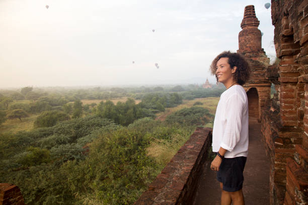 one young man, on top of temple, looking at view of Bagan, Myanmar Young traveller, wearing a white shirt and shorts, looking at some air got balloons and buddhist temples of Bagan archeological site, during golden hour. bagan archaeological zone stock pictures, royalty-free photos & images