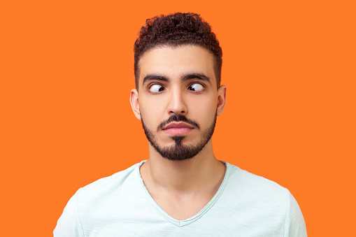 Portrait of comic positive brunette man with beard in casual white t-shirt looking cross-eyed, having fun with silly face expression, playing fool. indoor studio shot isolated on orange background