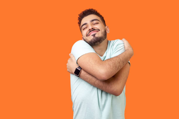 i love myself! portrait of egoistic brunette embracing himself and smiling form pleasure. indoor studio shot isolated on orange background - one man only imagens e fotografias de stock