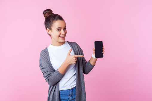 Look at mobile app! Portrait of positive teenage brunette girl with bun hairstyle in casual clothes pointing at cellphone and smiling joyfully at camera. studio shot isolated on pink background