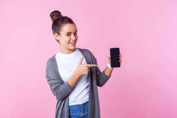 spójrz na aplikację mobilną! portret pozytywnej nastoletniej brunetki wskazującej na telefon i uśmiechniętej. różowe tło - telephone child isolated on white elegance zdjęcia i obrazy z banku zdjęć