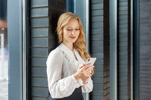 Portrait of stunning elegant businesswoman with long blond hair in white stylish shirt leaning on brick wall and smiling happily while using smartphone, reading message in social network. indoors