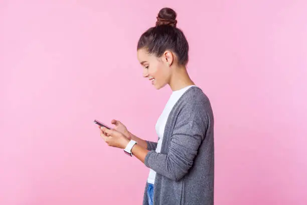 Photo of Side view of happy beautiful teenage girl using cell phone, feeling excited of chatting with friends. pink background