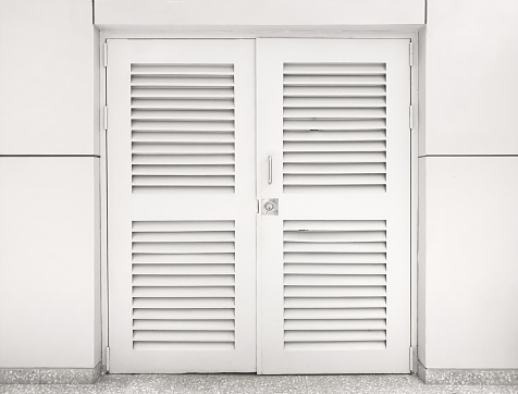 The louver wooden white door is locked with the white wall. Door with horizontal vents.  window with white shutters.